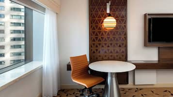 Habitación de hotel con un ventanal grande, una silla moderna, una mesita redonda pequeña, un televisor instalado en la pared y un suelo con diseño geométrico.