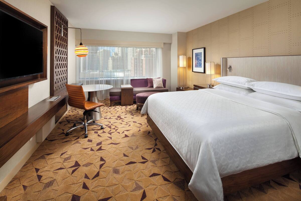 A hotel room with a large bed, a desk with a chair, a small table, a sofa, and a TV.