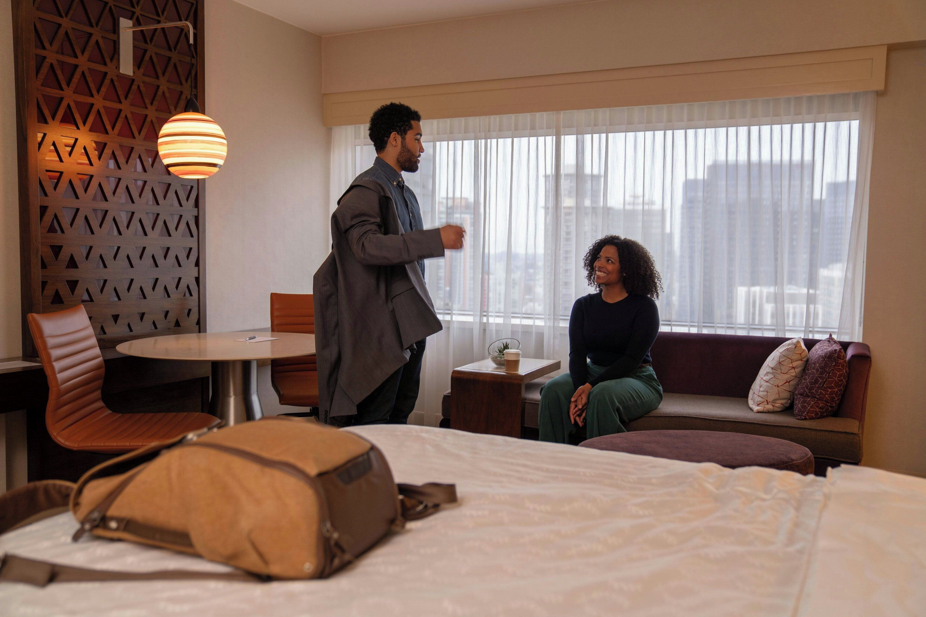 A hotel room with a bed, a seating area with a sofa and chairs, a small table, and a window with a city view.