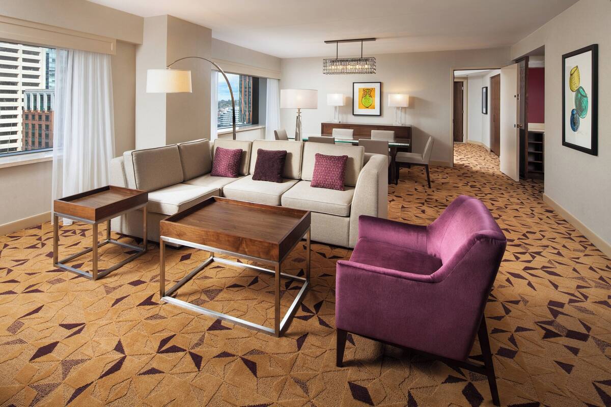 A hotel room with a sofa, armchair, coffee table, and dining area.