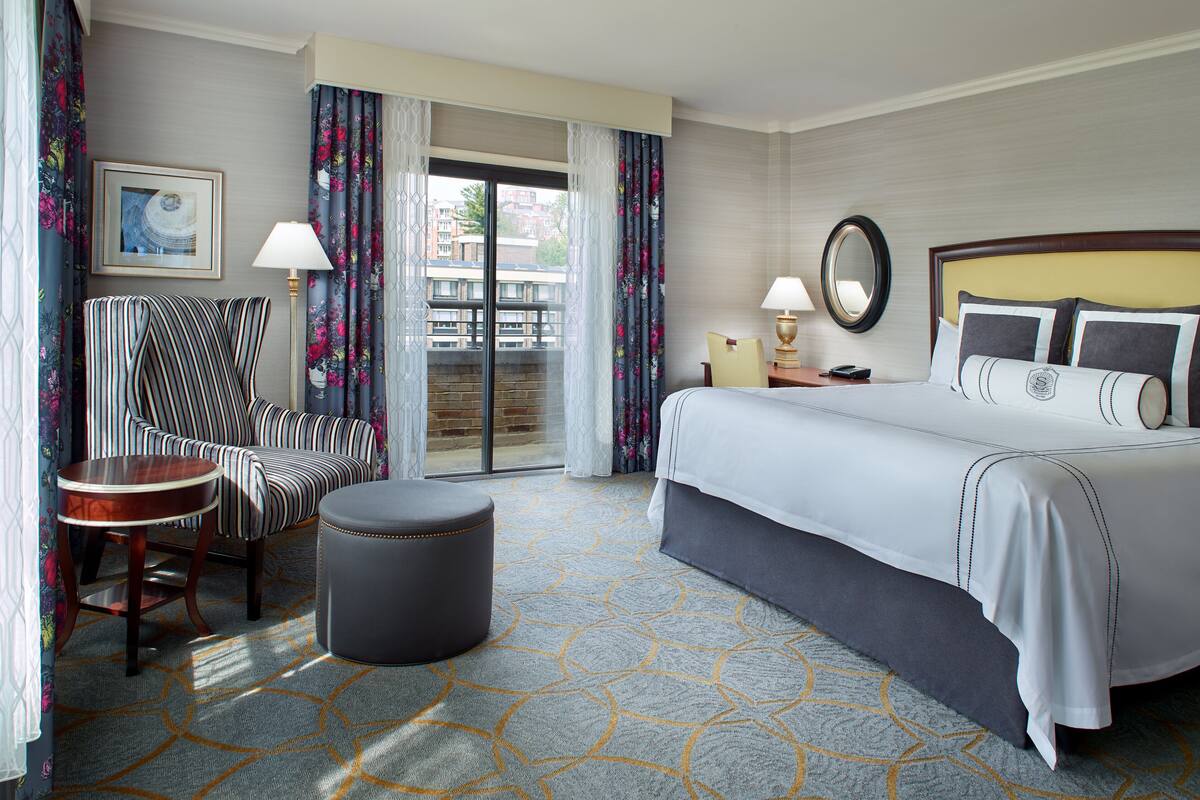 A hotel room with a bed, a chair, a table, a lamp, and a view of the outside.