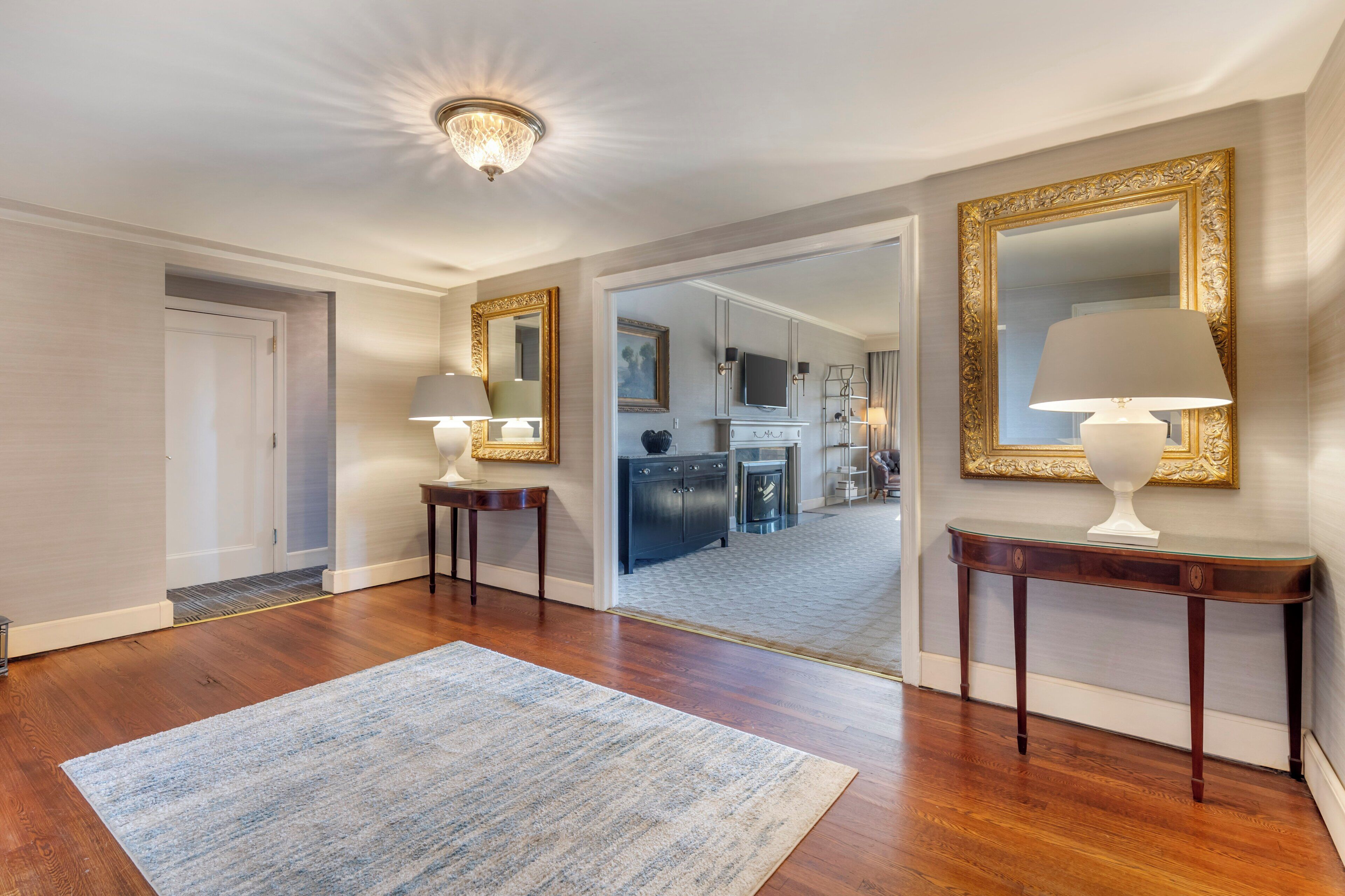 A spacious hallway with hardwood floors, a blue striped rug, and two ornate tables with lamps.