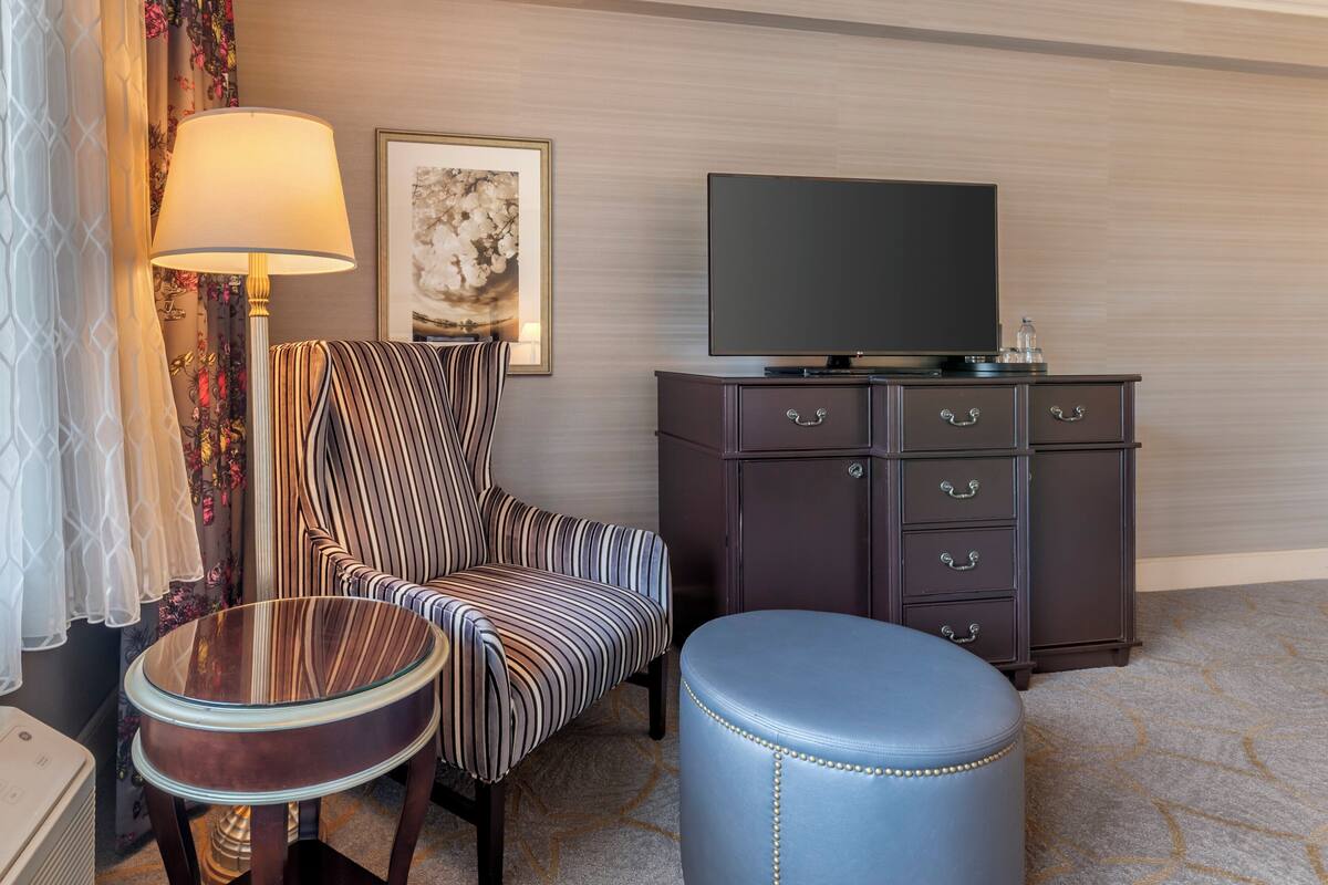 A hotel room with a striped armchair, a round glass table, a dark wooden cabinet with drawers, a television on top, and a blue ottoman.