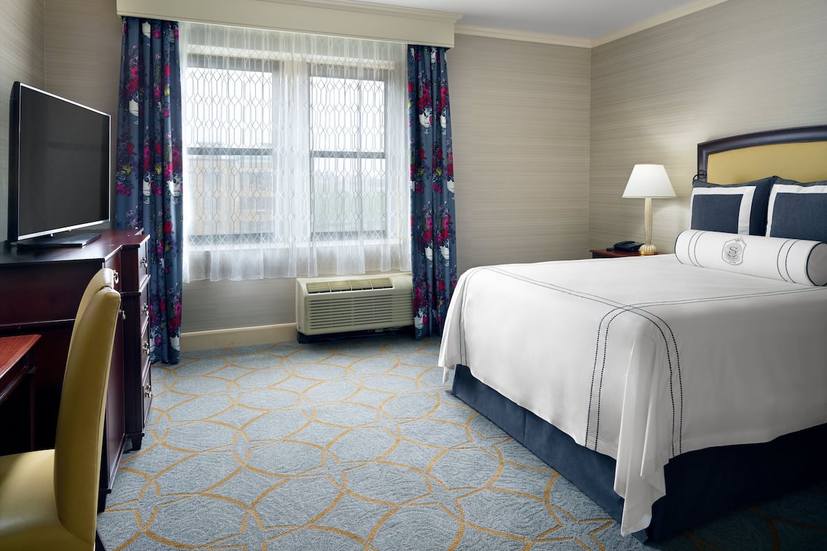 A hotel room with a bed, a desk with a television, a chair, a nightstand with a lamp, and a window with curtains.