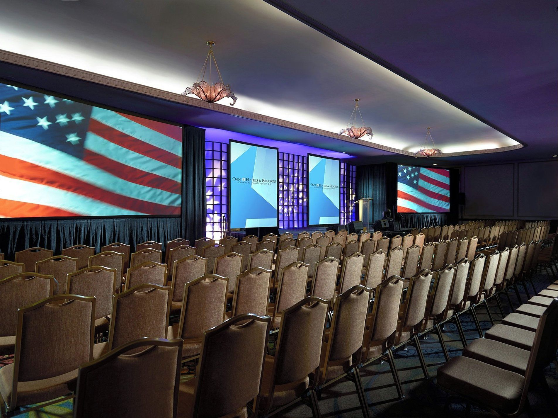 A conference room with rows of chairs, a stage with a screen displaying the hotel name, and American flags on the walls.