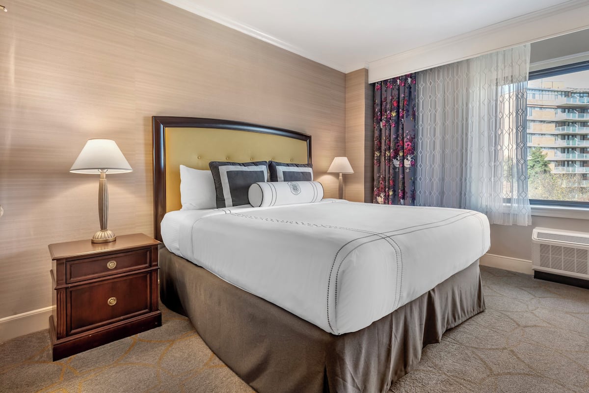 A hotel room with a large bed, two bedside tables with lamps, a window with curtains, and a view of buildings outside.