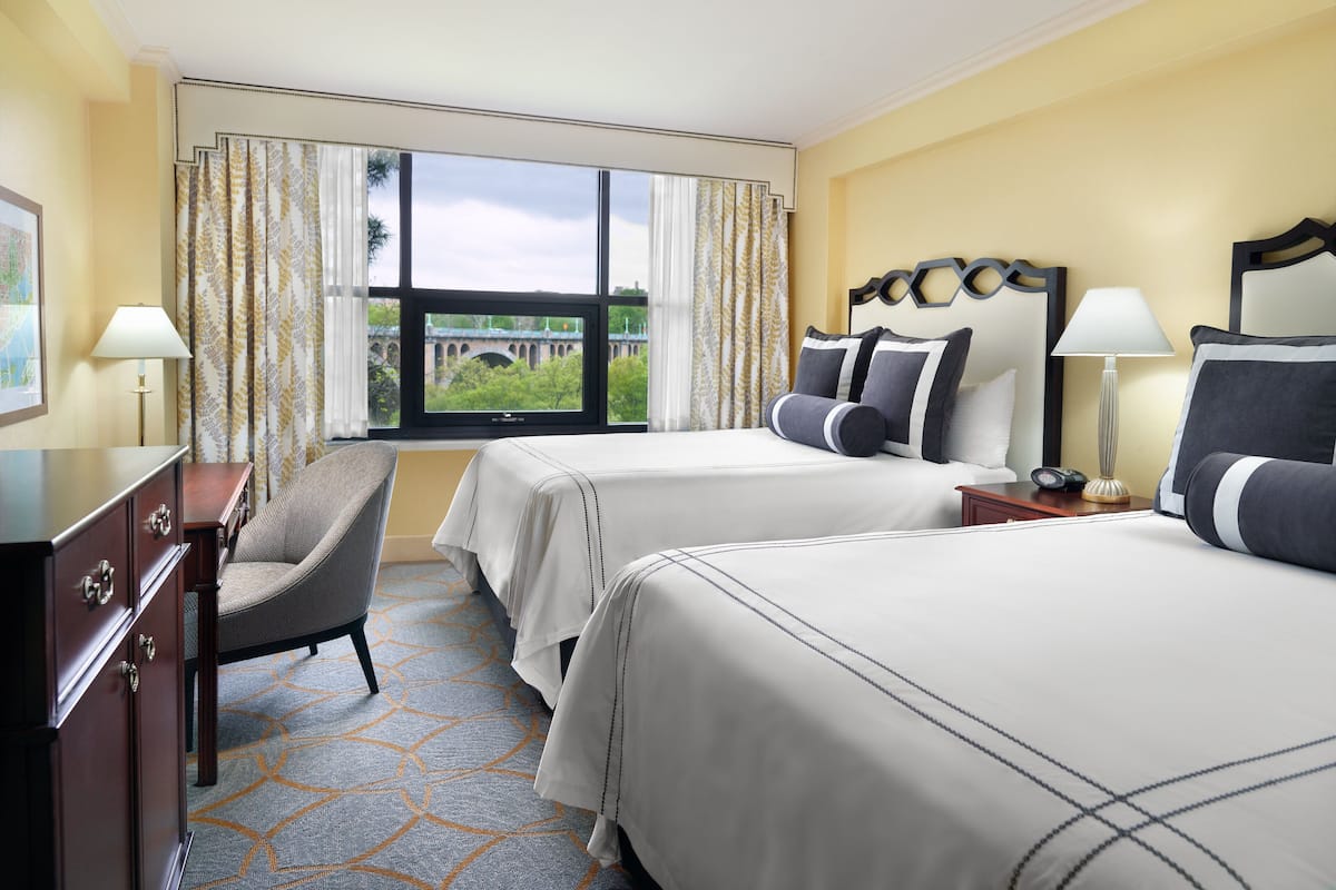 A hotel room with two beds, a desk, a chair, and a large window with a view of a bridge.