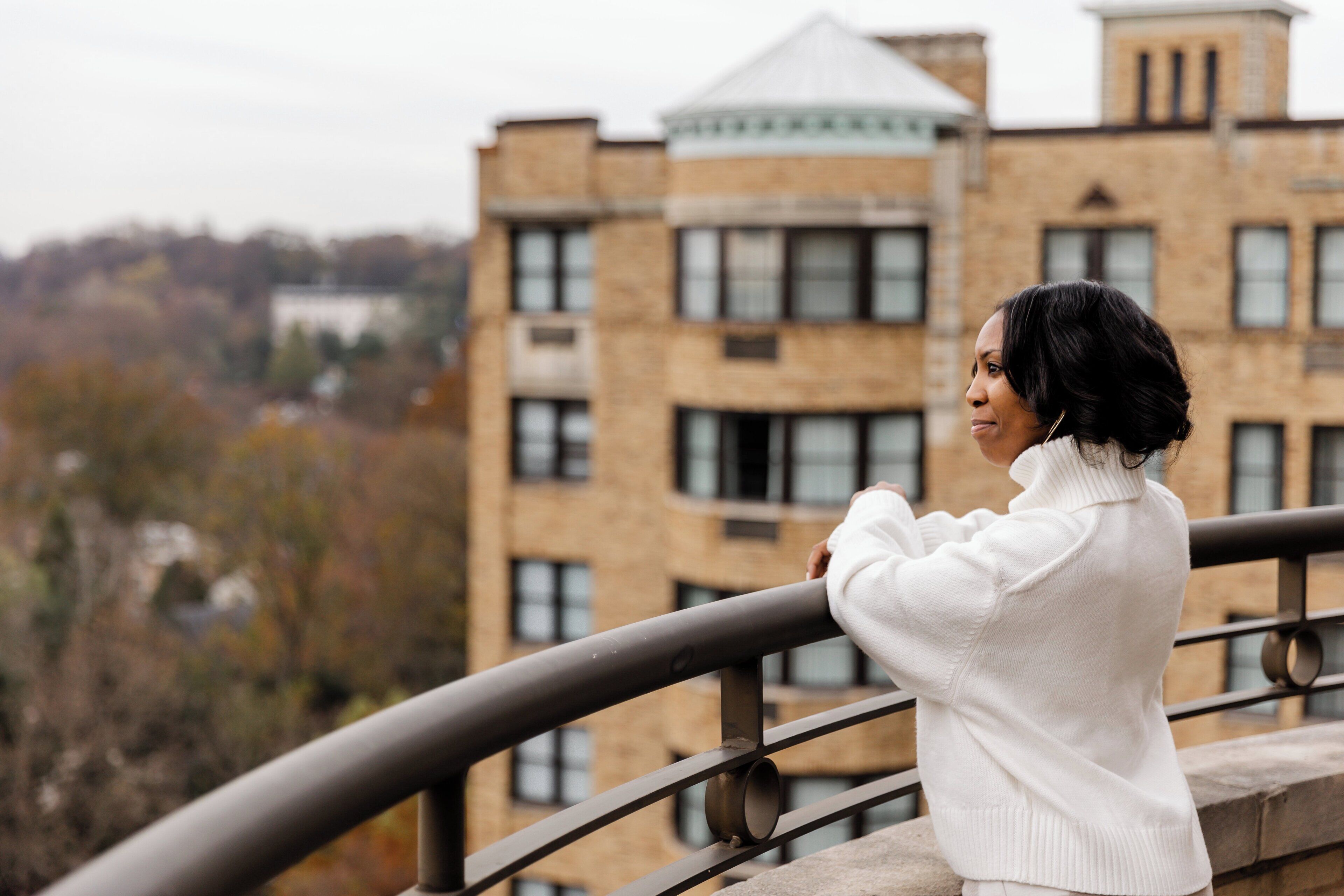 A person standing on a balcony with a view of a brick building and trees.