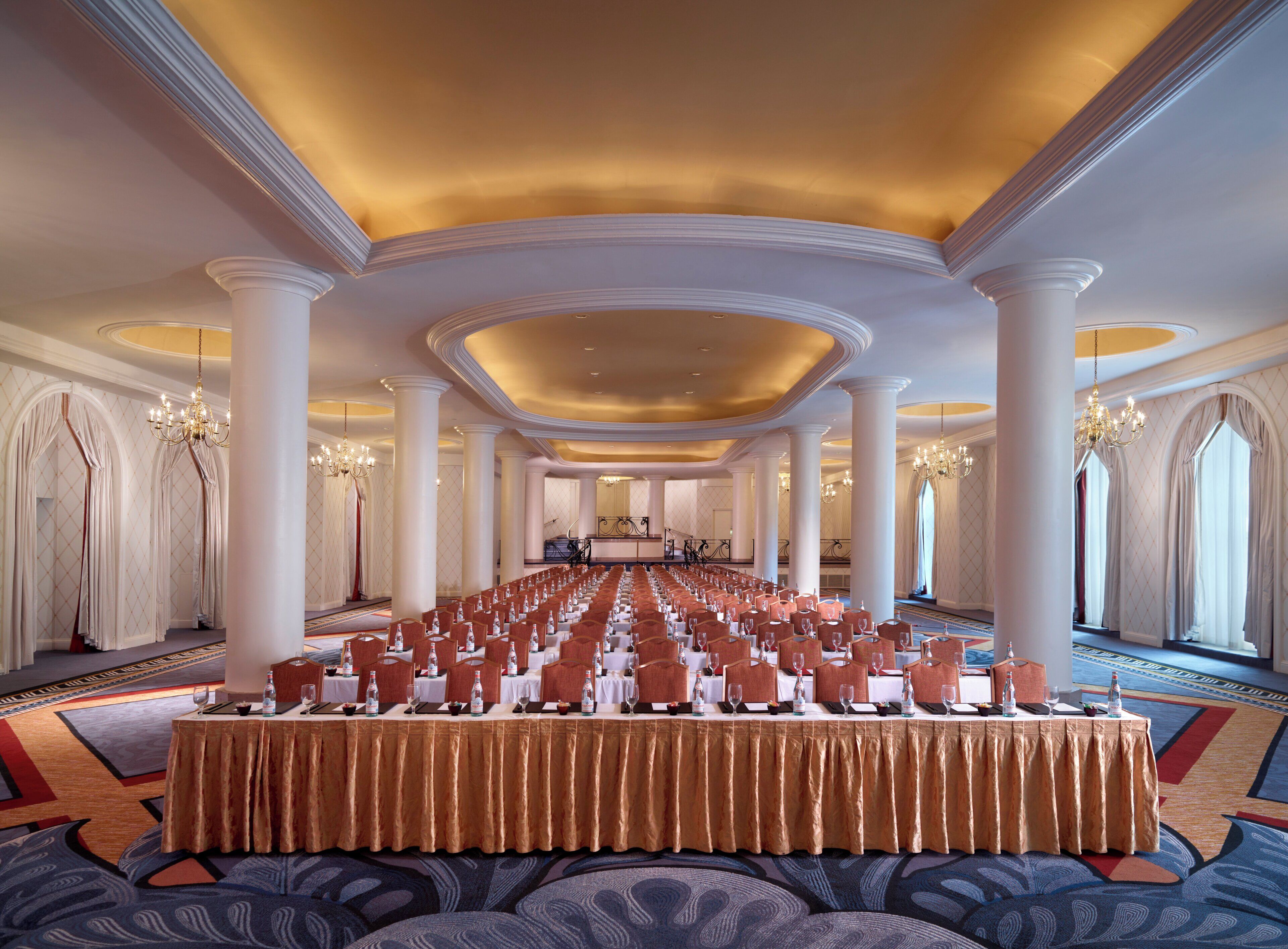 A large banquet hall with rows of tables set for a formal event, featuring a golden tablecloth, white chairs, and chandeliers.