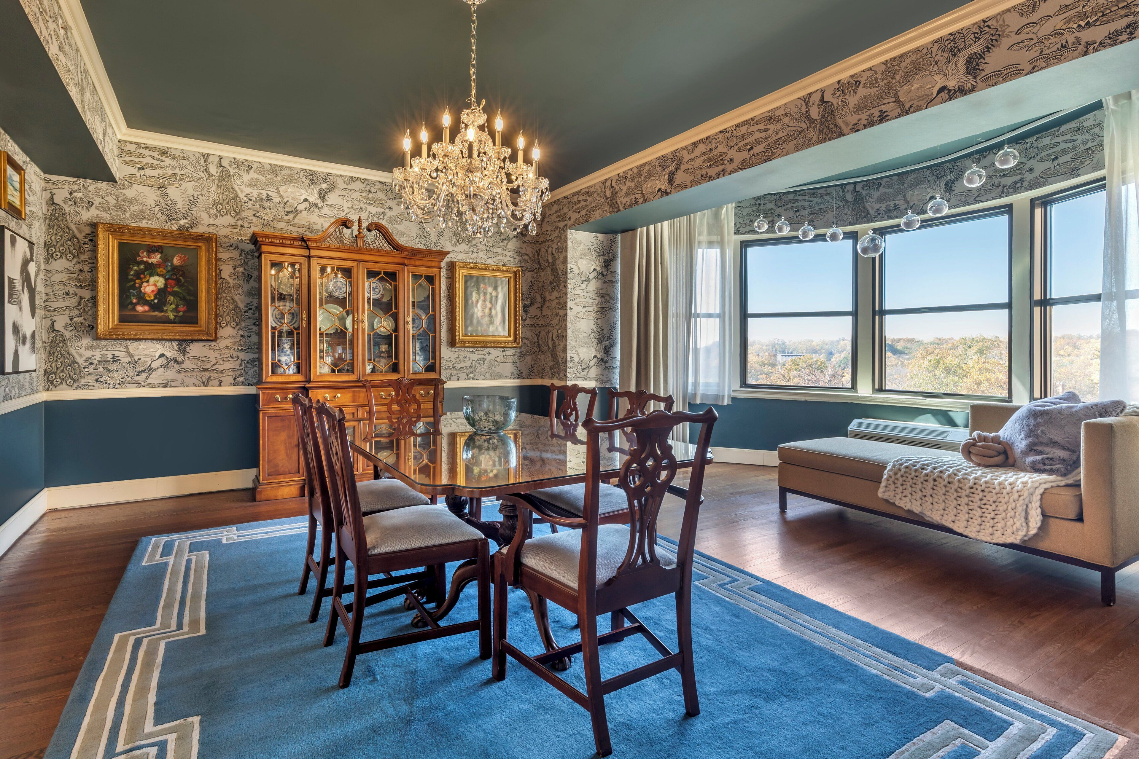 A dining room with a large table, chairs, a chandelier, and a view of the outdoors through large windows.