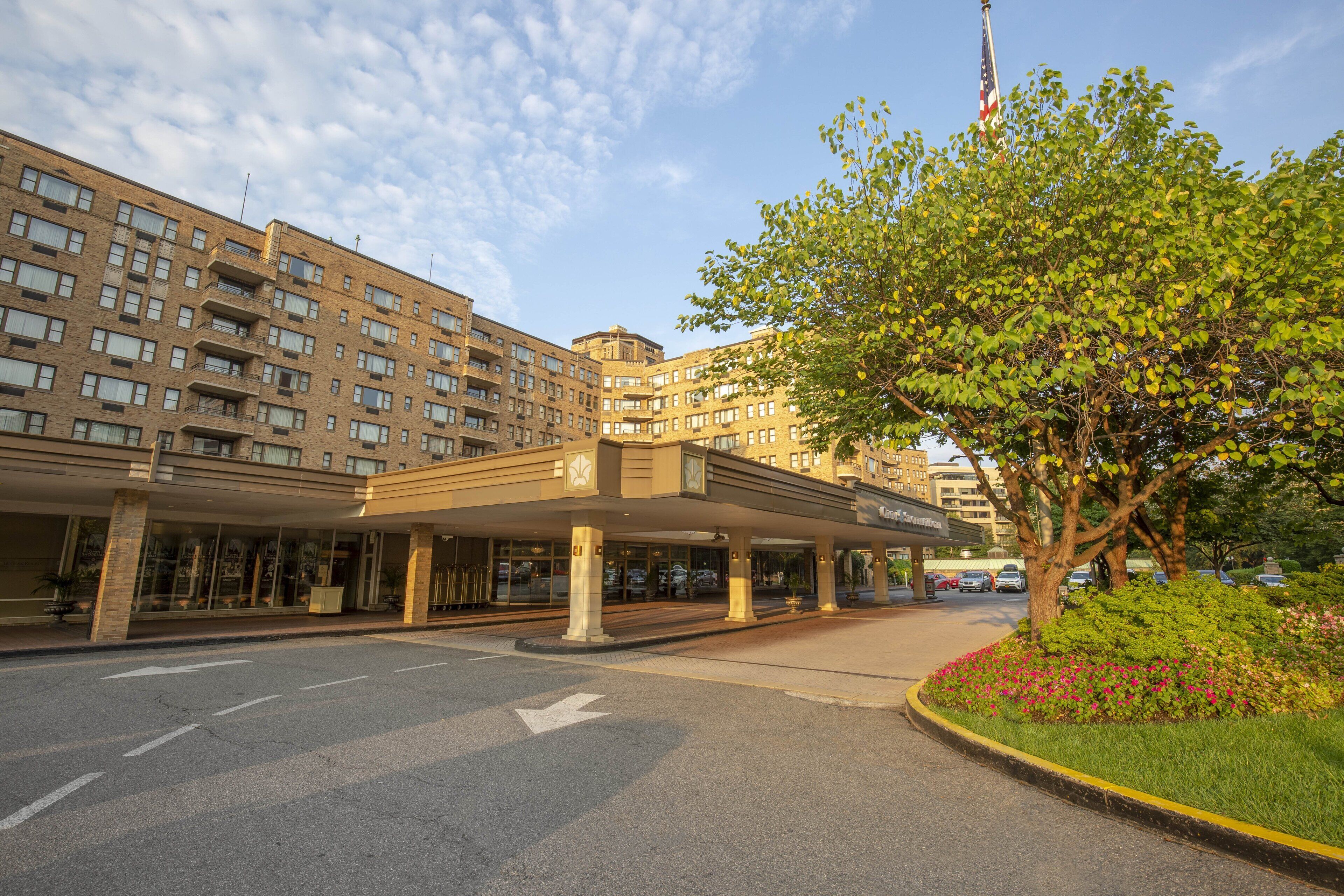 A multi-story hotel with a covered entrance, a flagpole with an American flag, and a landscaped area with flowers.