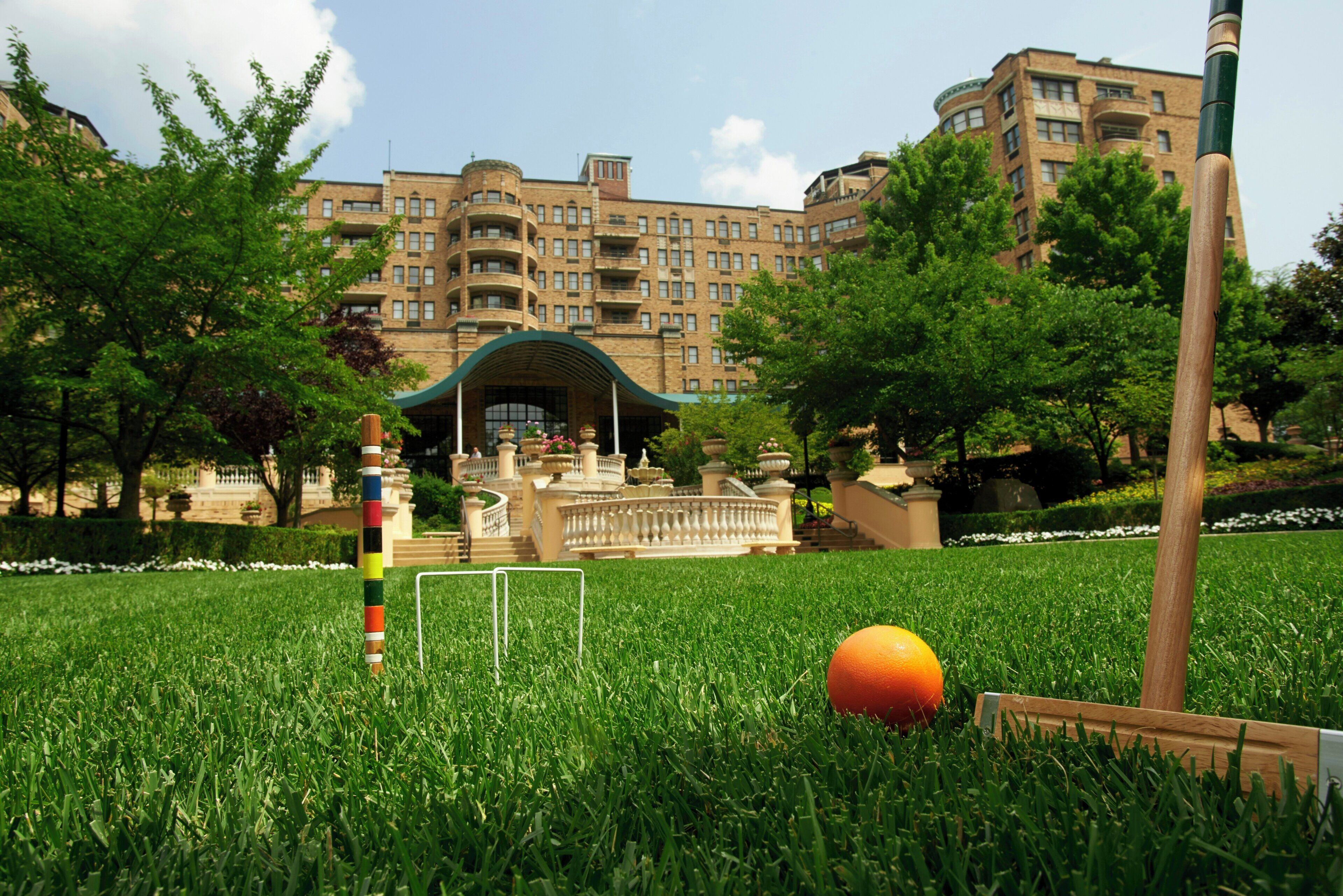 Un césped verde con una pelota de croquet y mazos, un poste colorido, y un edificio con varios pisos y balcones al fondo.