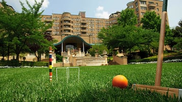 Un césped verde con una pelota de croquet y mazos, un poste colorido, y un edificio con varios pisos y balcones al fondo.