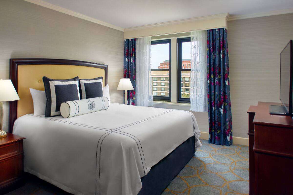 A hotel room with a large bed, two bedside lamps, a wooden nightstand, a dresser, and a television on a wooden stand.