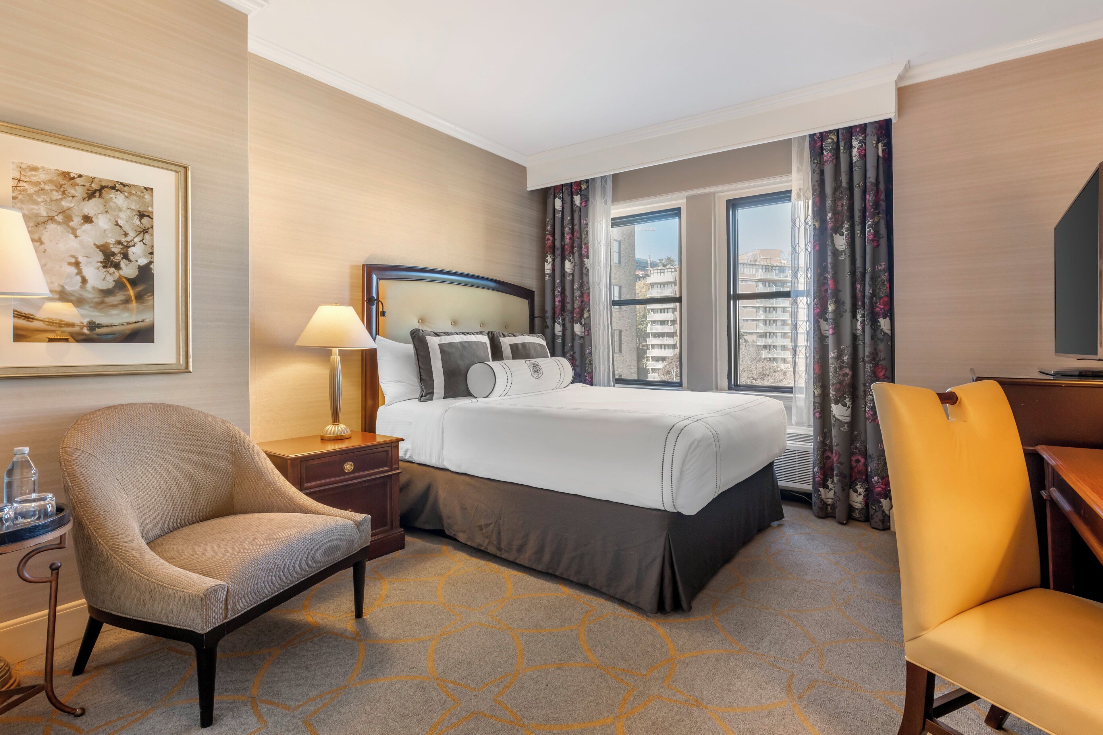A hotel room with a bed, two chairs, a desk, a lamp, a framed picture, and a window with curtains.