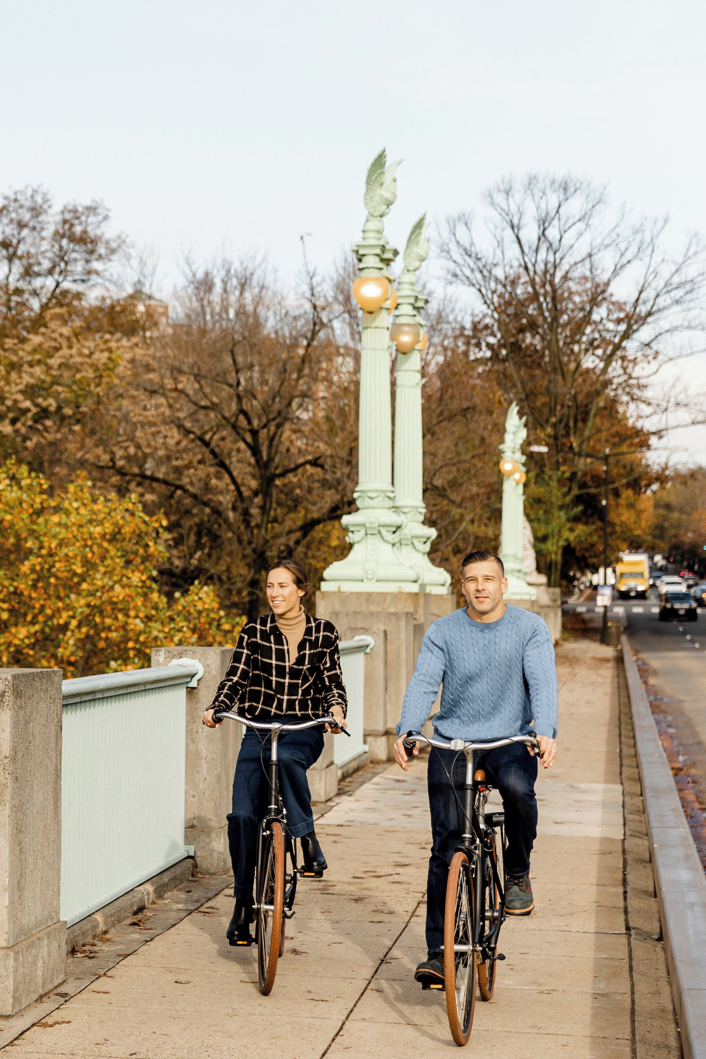 Dos personas andando en bicicleta sobre un puente con columnas decorativas y árboles otoñales al fondo.
