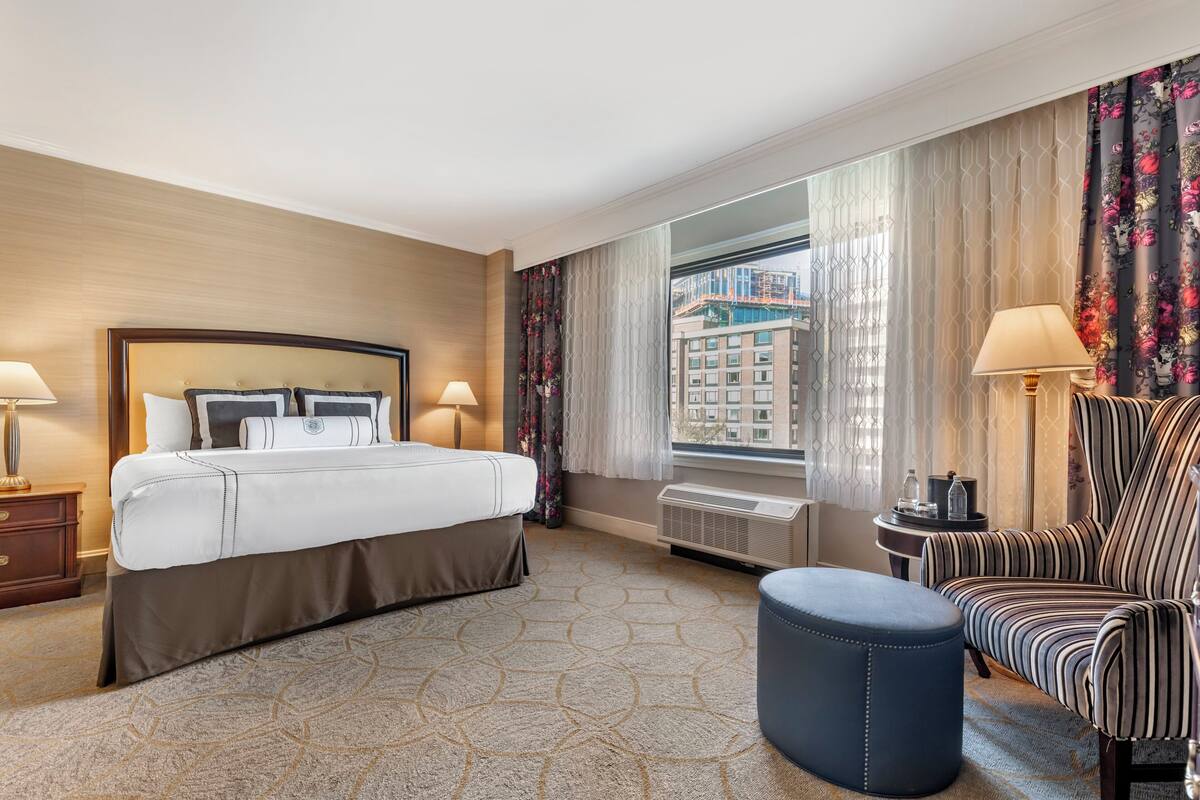 A hotel room with a large bed, a chair, a small table, and a view of a building through the window.