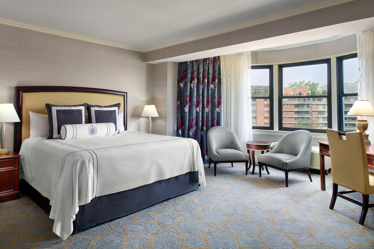 A hotel room with a large bed, two chairs, a desk, and a view of buildings through the windows.