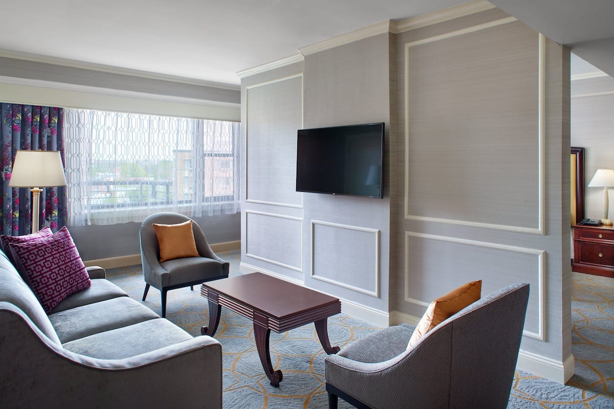 A hotel room with a grey sofa, a matching armchair, a small wooden table, and a flat-screen TV mounted on the wall.