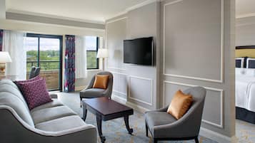 A hotel room with a grey sofa, armchairs, a small table, a TV mounted on the wall, and a large window with curtains.