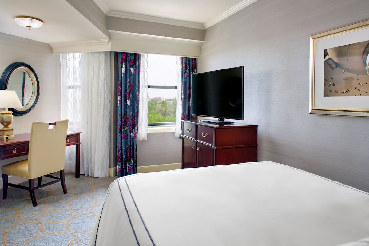 A hotel room with a bed, a desk with a chair, a television, and a window with curtains.