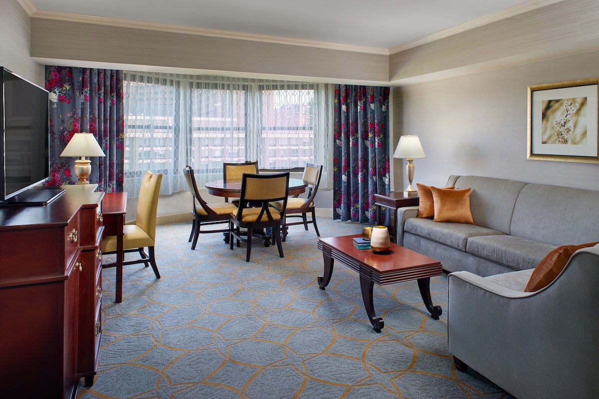 A hotel room with a dining area, a sofa, a TV, and a painting on the wall.