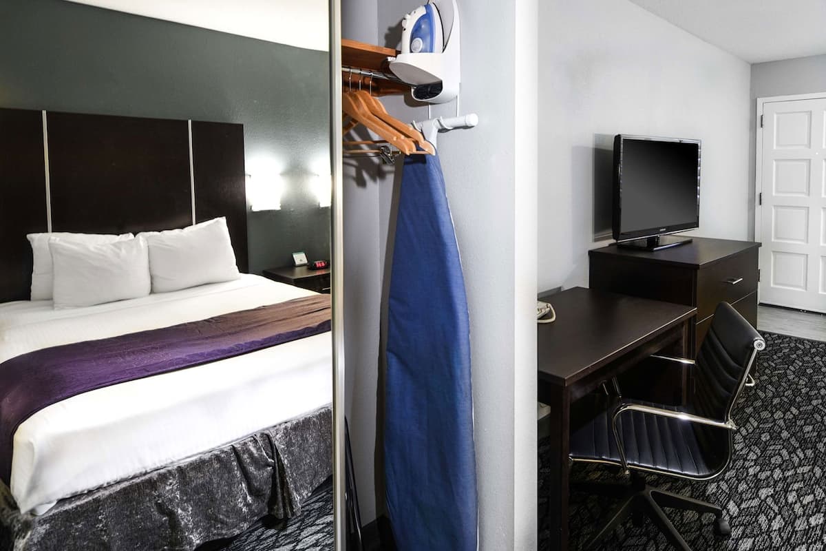 Ein Hotelzimmer mit einem Bett, einem Schreibtisch mit Fernseher, einem Sessel und einem Kleiderständer mit einem blauen Kleidungsstück.