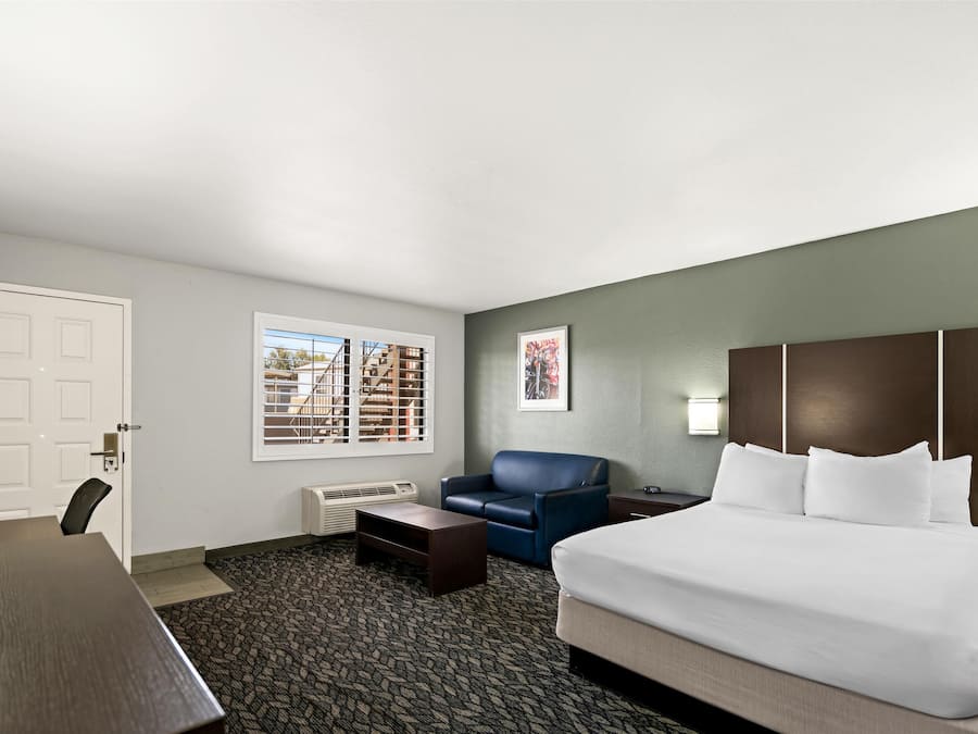 A hotel room with a bed, a desk, a chair, a sofa, a window with blinds, and a framed picture on the wall.