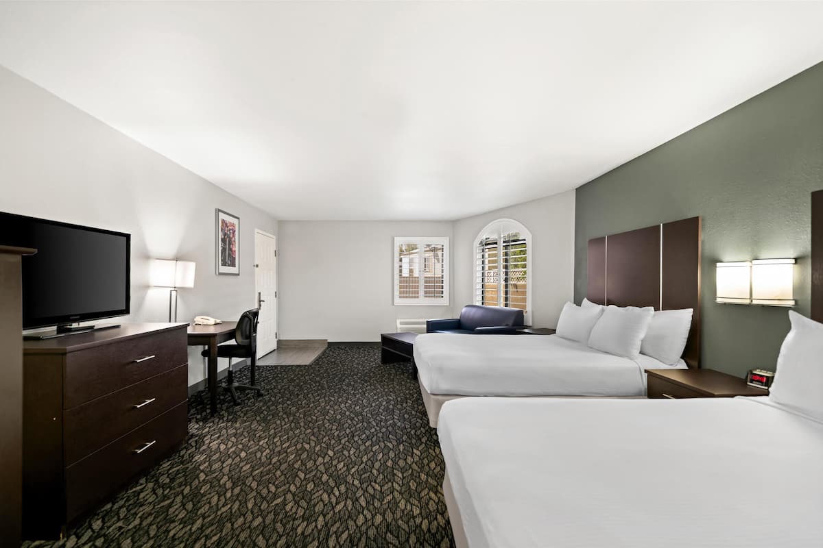 Ein Hotelzimmer mit zwei Betten, einem Schreibtisch mit Stuhl, einem Fernseher, einer Lampe und einem Fenster mit Jalousien.