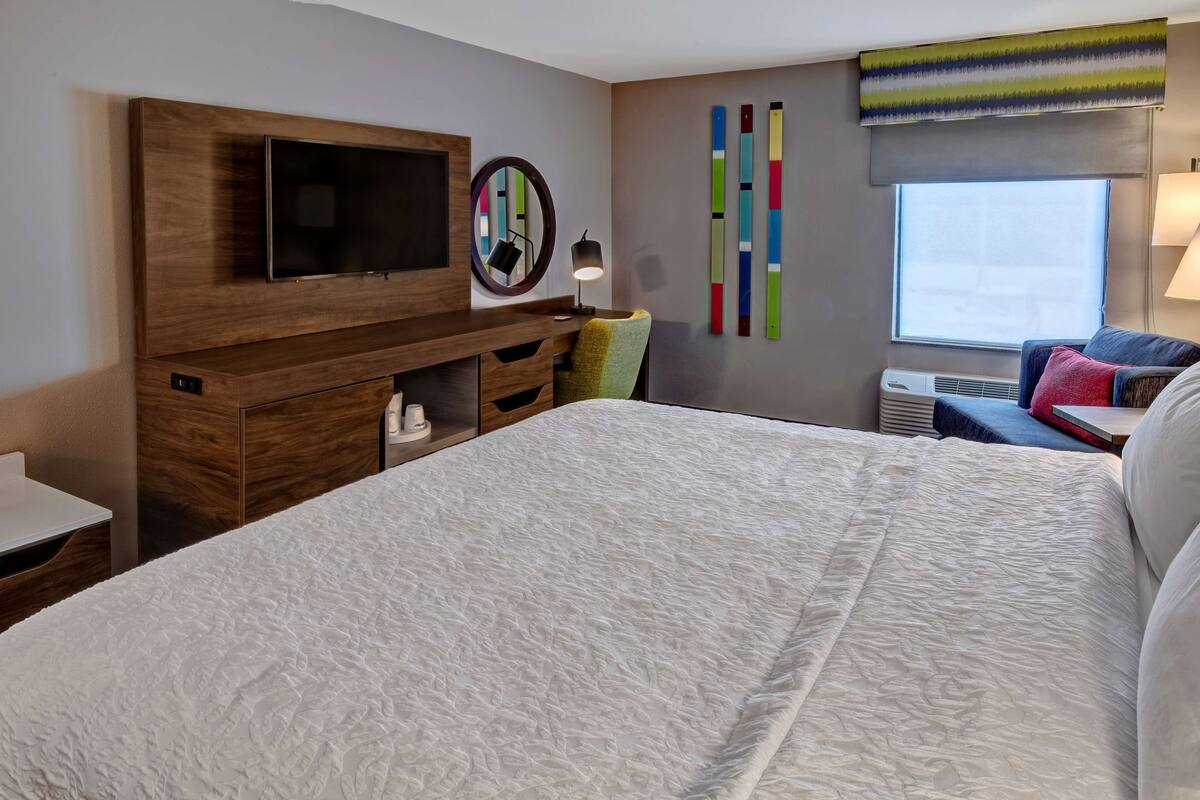 Habitación de hotel con una cama grande, un televisor montado en un mueble de madera, un espejo redondo, una lámpara, un adorno de pared colorido y una ventana con aire acondicionado.