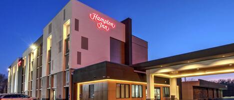 A Hampton Inn hotel with a modern design, featuring a neon sign, a covered parking area, and a well-lit entrance.