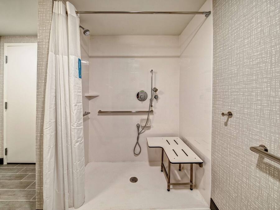 Eine begehbare Dusche mit weißer Duschvorhang, einer Handbrause, einer Haltevorrichtung und einer kleinen Sitzbank.
