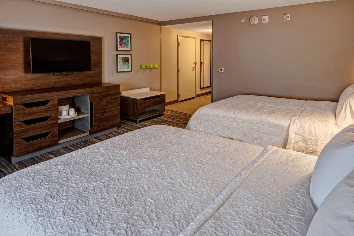 Habitación de hotel con dos camas, un televisor montado en un mueble de madera, una zona de baño pequeña y una puerta que da a otra habitación.