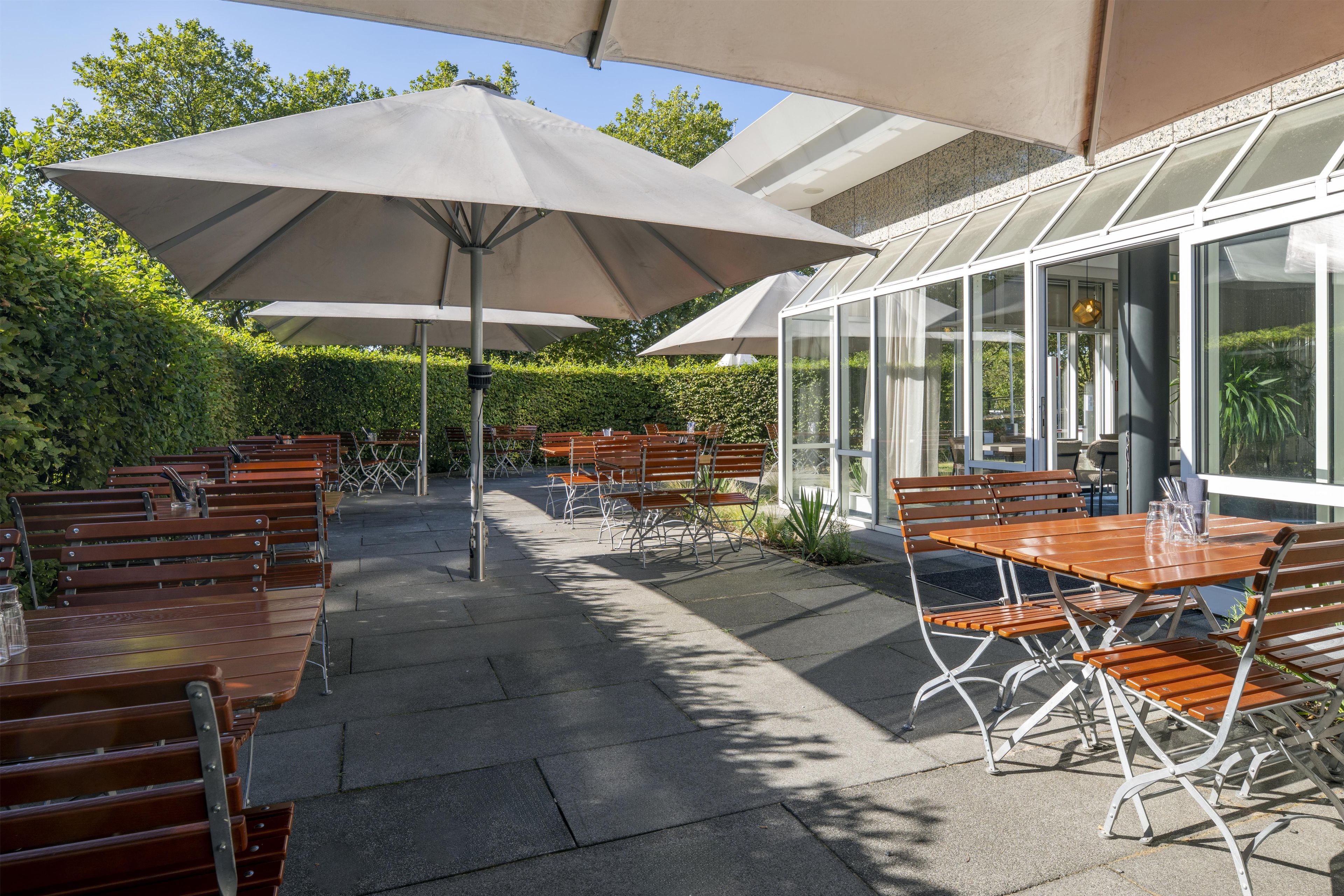 An outdoor patio with wooden tables and benches, covered by large umbrellas, and a glass conservatory adjacent to the seating area.