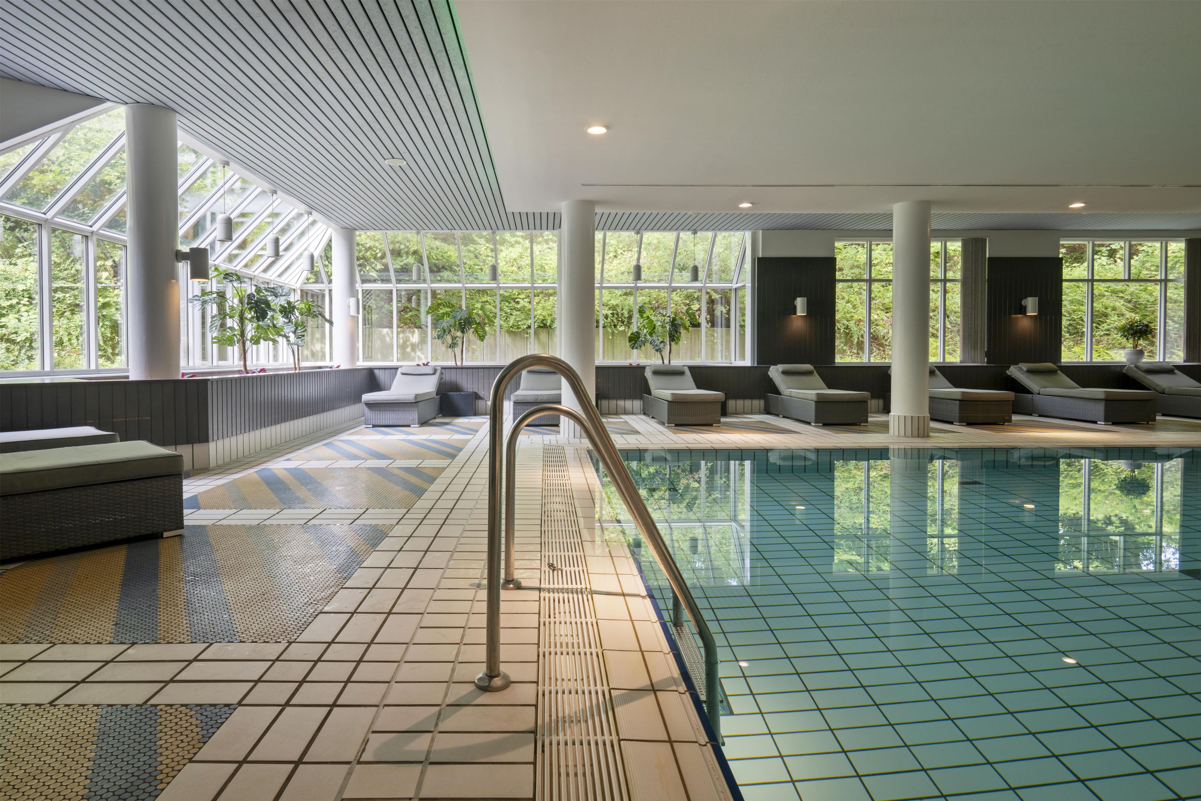 Ein Swimmingpool mit Fliesenboden, Liegestühlen und großen Fenstern mit Blick auf Grünflächen.