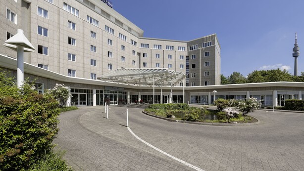 Un hotel moderno con entrada cubierta, varios pisos y una torre destacada al fondo.