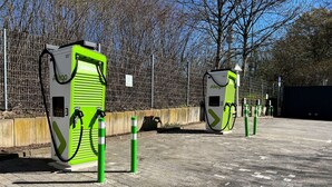 Dos estaciones de carga para vehículos eléctricos verdes con un área de estacionamiento pavimentada y una cerca al fondo.