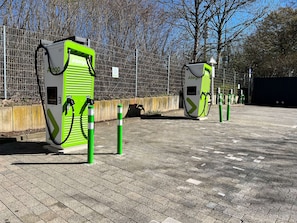 Two green electric vehicle charging stations with a paved parking area and a fence in the background.
