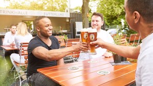 Un grupo de personas brindando con vasos de cerveza en una mesa al aire libre con superficie de madera y posavasos.