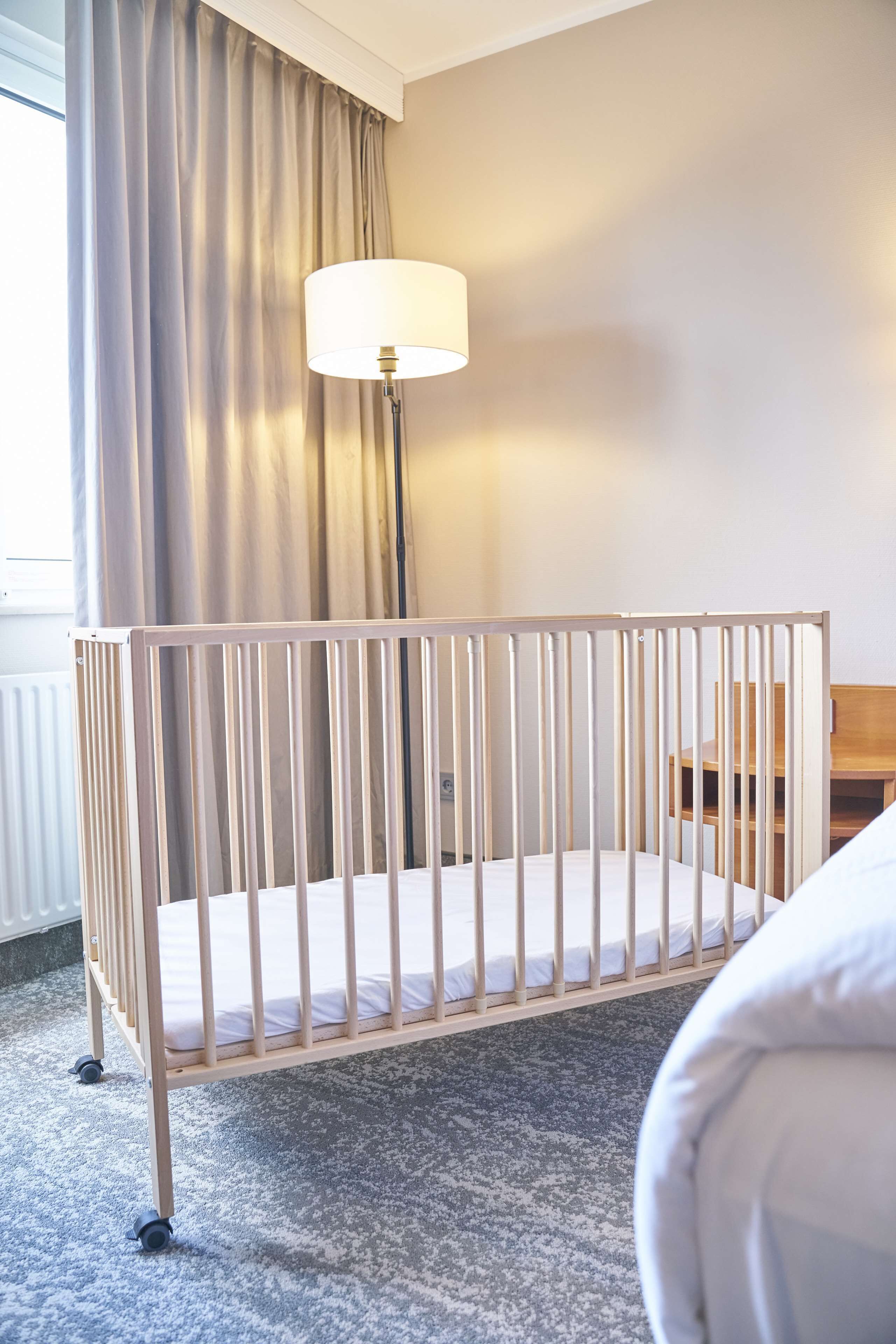 A baby crib with a white mattress, a floor lamp, and a wooden nightstand in a hotel room.