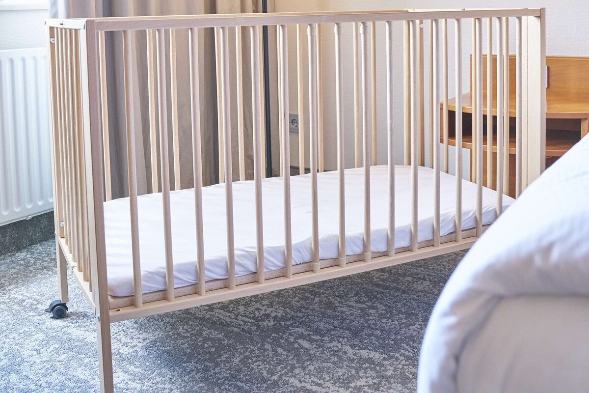 A baby crib with a white mattress, a floor lamp, and a wooden nightstand in a hotel room.