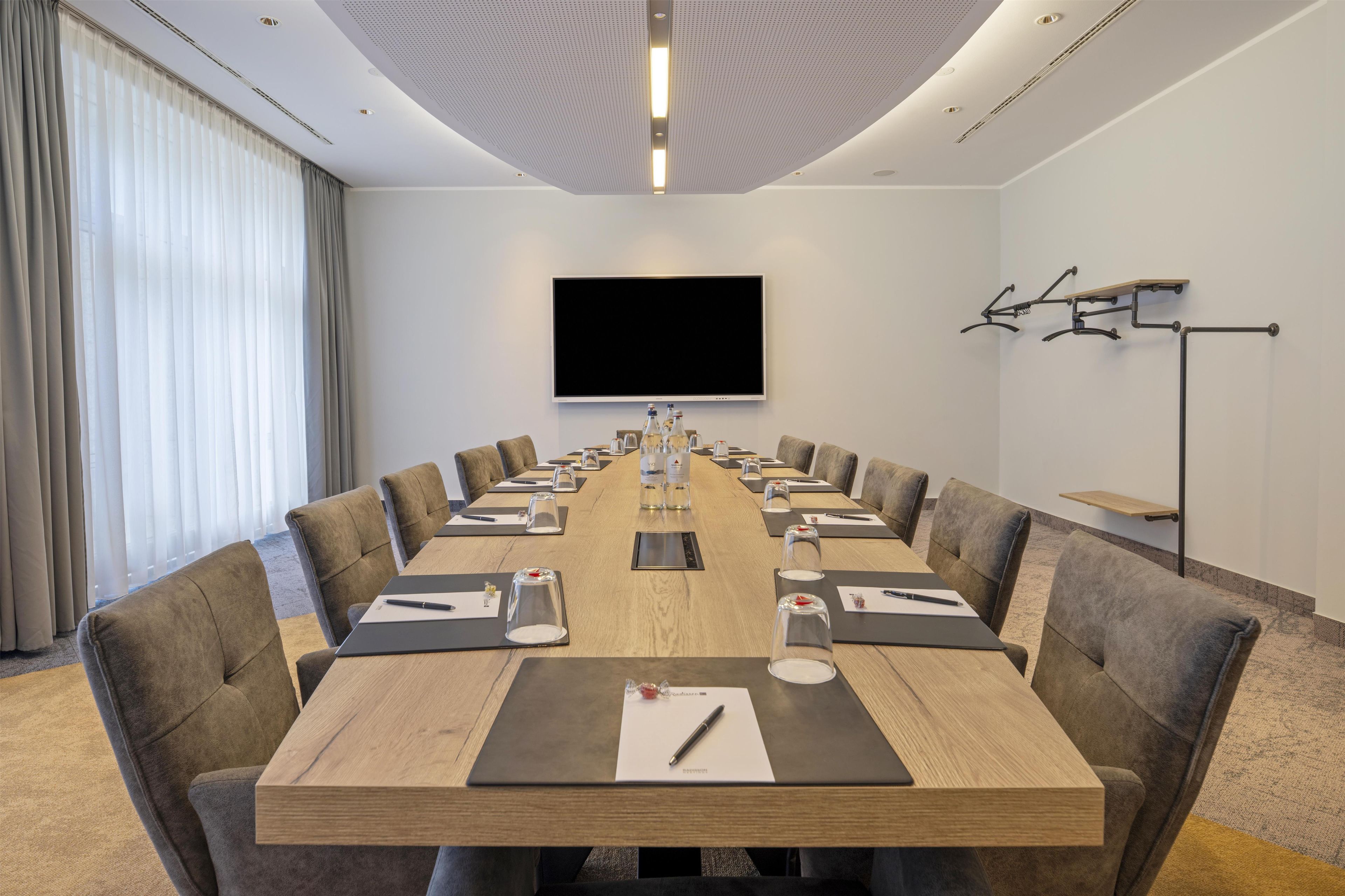 Een vergaderzaal met een lange tafel, stoelen, een flatscreen-tv en een beamerstandaard.