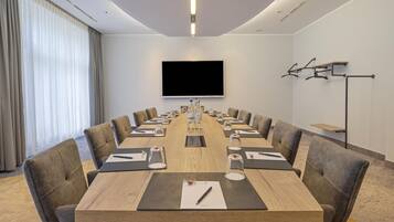 Een vergaderzaal met een lange tafel, stoelen, een flatscreen-tv en een beamerstandaard.