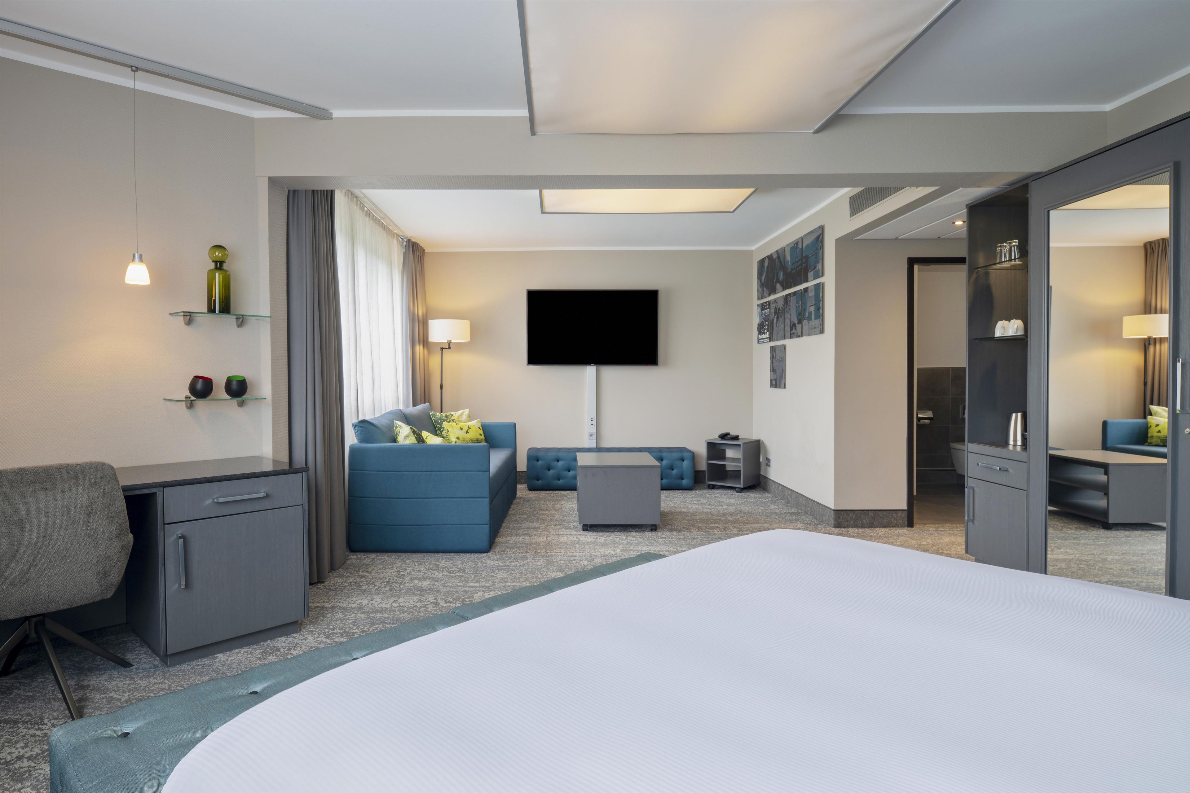 Habitación de hotel con una cama grande, un escritorio, un sofá, un televisor y un baño visible a través de una puerta abierta.