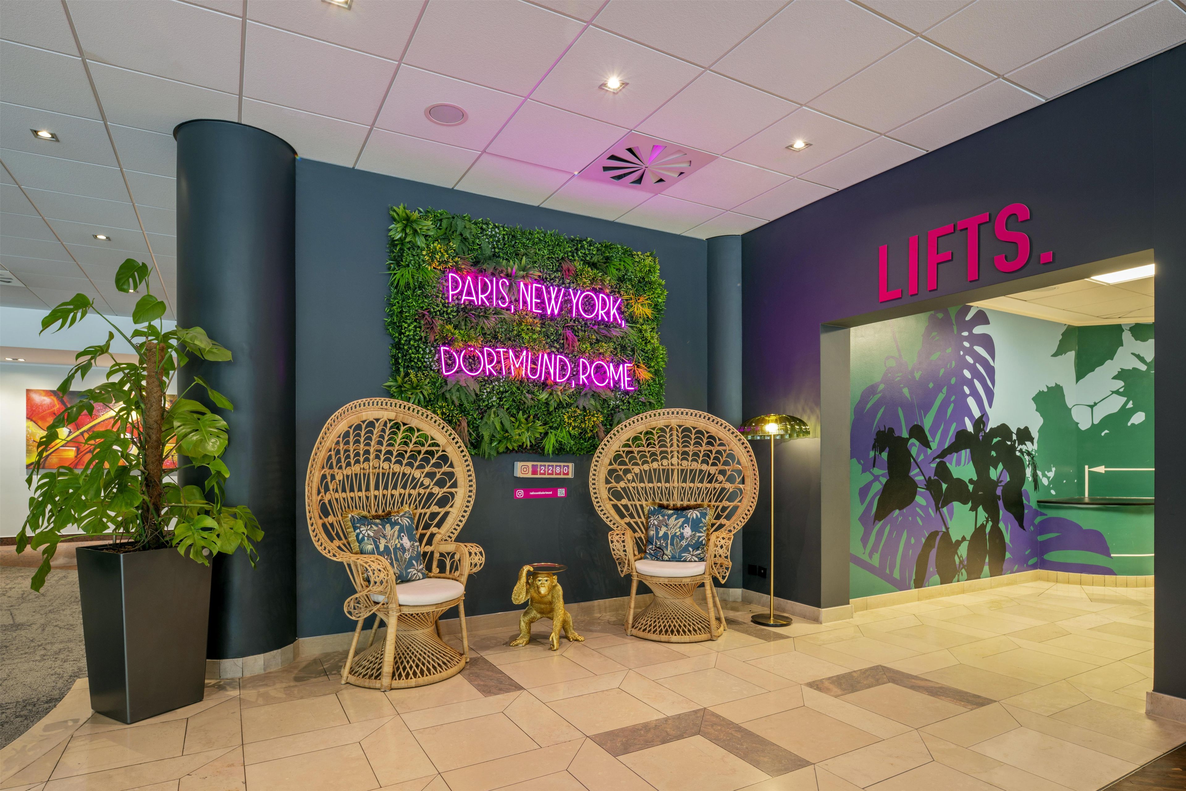A lobby with two wicker chairs, a neon sign, and a wall with a green leafy design.