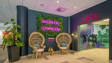 A lobby with two wicker chairs, a neon sign, and a wall with a green leafy design.