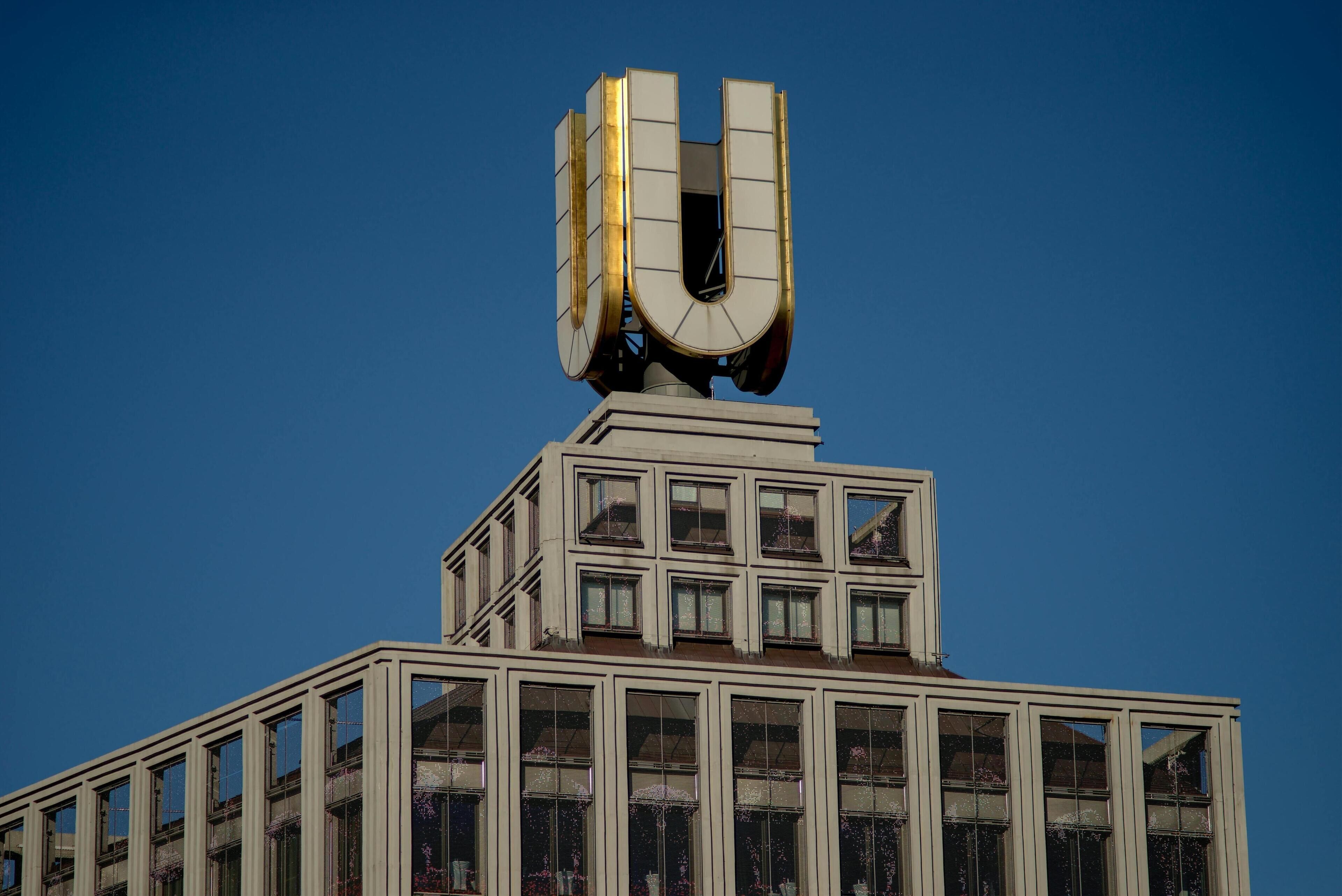 A building with a prominent U-shaped structure on top, large windows, and a clear blue sky in the background.