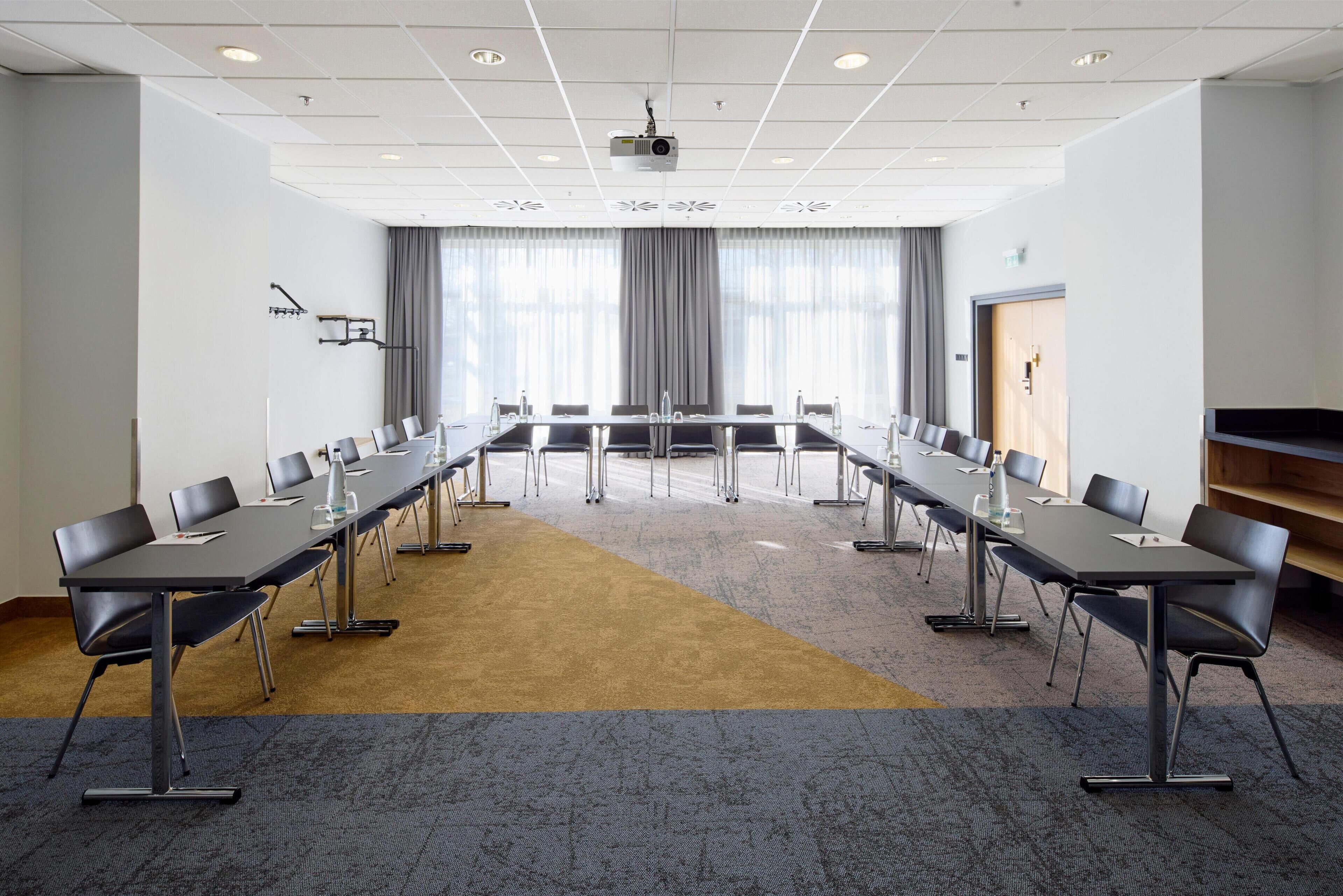 Ein Konferenzraum mit langem Tisch, Stühlen, einem Projektor und einem großen Fenster mit Vorhängen.