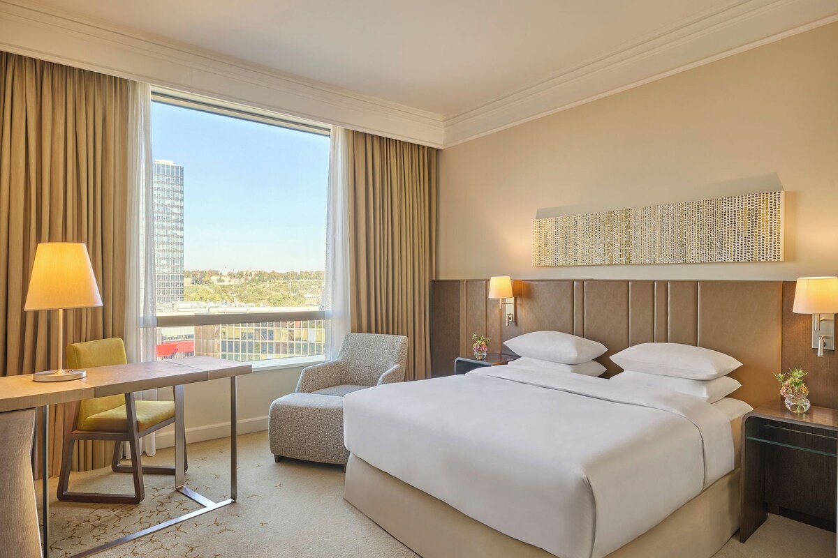 A hotel room with a bed, a desk, a chair, a nightstand, and a view of a cityscape.