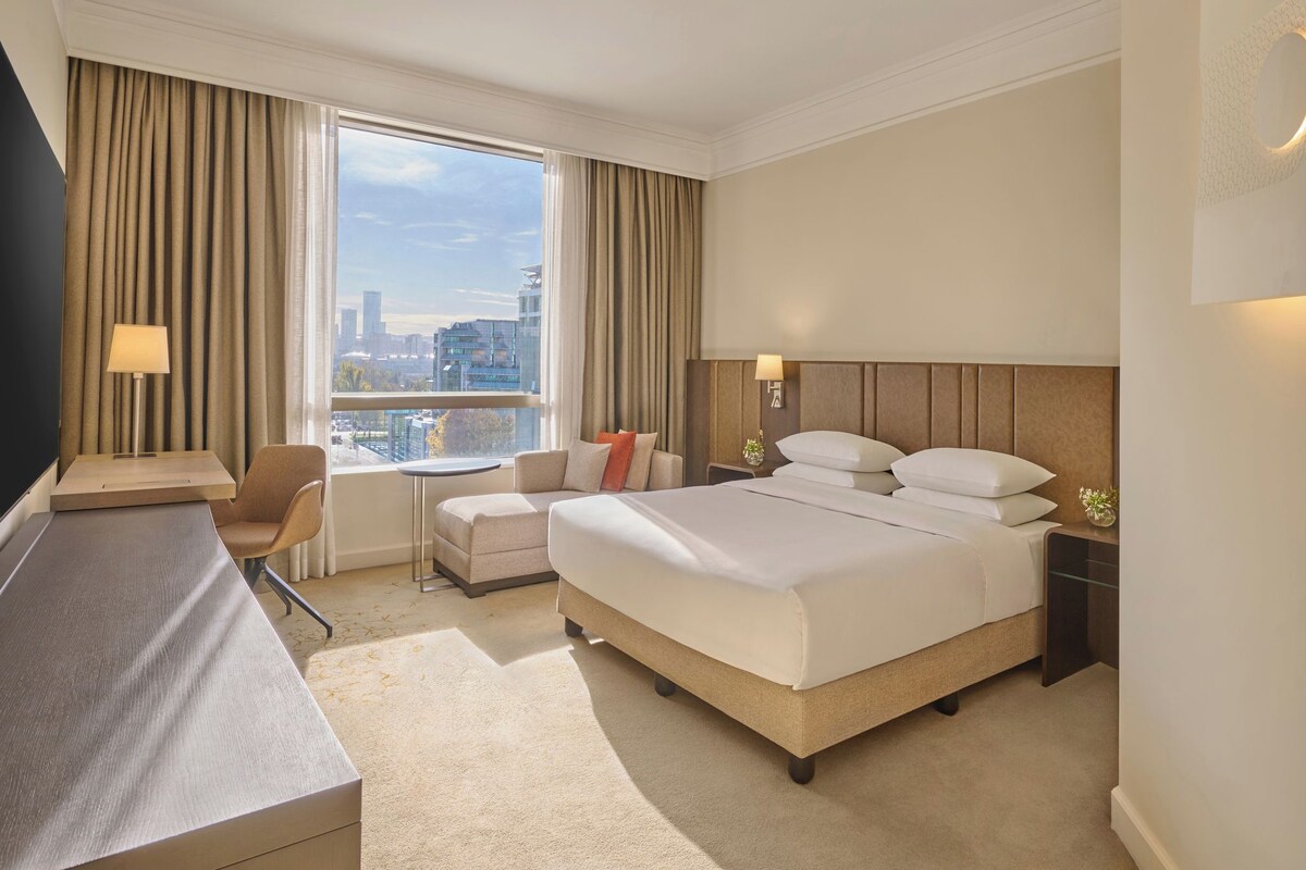 A hotel room with a large bed, a desk, a chair, a small table, and a view of the city.